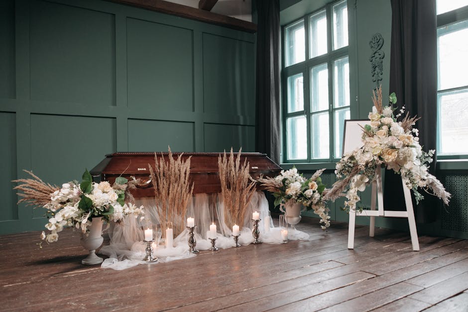 A floral arrangement featuring predominantly white flowers, including roses and lilies, arranged in two large white vases on either side of a white easel displaying a white floral tribute. The flowers