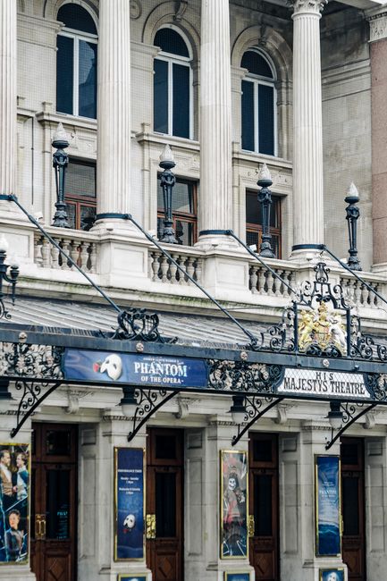 The image features the ornate façade of the Majesty's Theatre, showcasing classical architectural details such as tall arched windows with decorative molding, stone balconies with balustrades, and in
