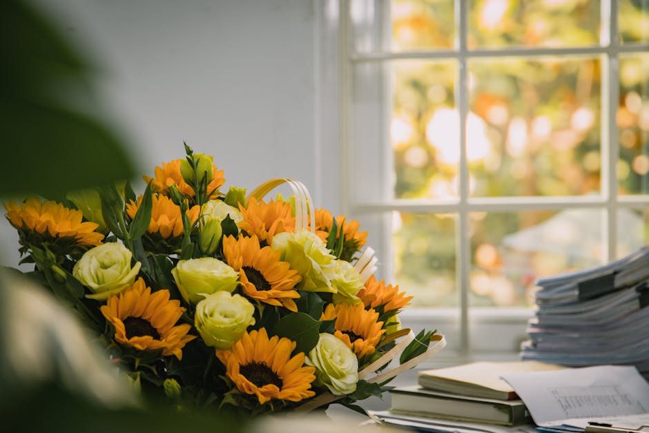 A close-up of a vibrant floral arrangement featuring bright yellow sunflowers and cream-coloured roses with green foliage, placed on a desk near a window. The flowers are fresh, with the petals and st