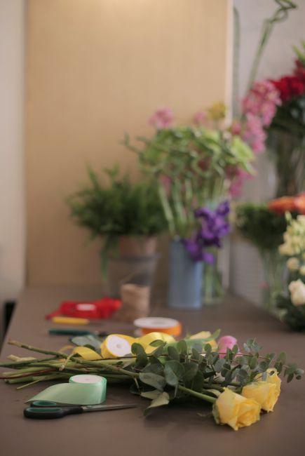 A close-up of fresh yellow roses with green foliage lying on a grey work surface, surrounded by floral arranging tools such as scissors, floral tape, and ribbon. In the background, various flower arra