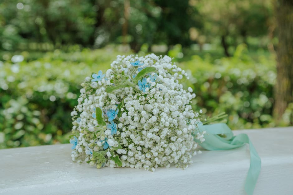 A bouquet arrangement of white gypsophila (baby's breath) with small blue forget-me-not flowers interspersed throughout, held together with a light green ribbon. The bouquet rests on a white wooden su