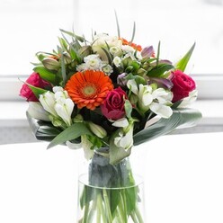 This expertly curated Bexleyheath bouquet is a visual symphony, designed to stir the senses and warm the heart. Creamy peach gerberas form a gentle halo, their petals diffusing light like the early morning sun rising over Bexleyheath's tree-lined avenues. At their core, brilliant orange gerberas burn with festive energy, drawing the eye amid the lush composition. Bold fuchsia roses lend a layer of opulent depth-each tightly furled bloom suggesting passion and elegance-while clusters of ivory alstroemeria and dainty white spray roses add brightness and gentle textural contrast. The abundance of foliage is as practical as it is poetic: paddle-shaped, glossy leaves curve gracefully around the bouquet's base, framing and protecting the arrangement in a manner reminiscent of the careful attention paid by local market florists. Light filters through from behind, illuminating each petal in a celebration of colour that feels fresh and optimistic. Hints of subtle fragrance-a blend of dewy roses, light lilies, and freshly snipped stems-suggest a bouquet ready for joyful milestones, brunch at a friend's on Oaklands Road, or expressing gratitude. This creation, rooted in the community and shaped by skilled hands, invites new beginnings and treasured moments.